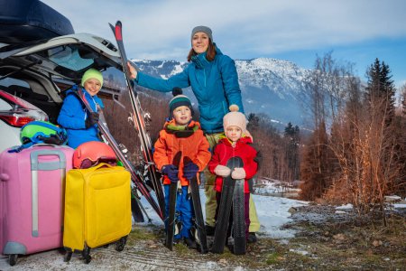 Photo for Mother stand holding ski by open car trunk arrived with three children at alpine skiing resort unloading suitcases and baggage - Royalty Free Image