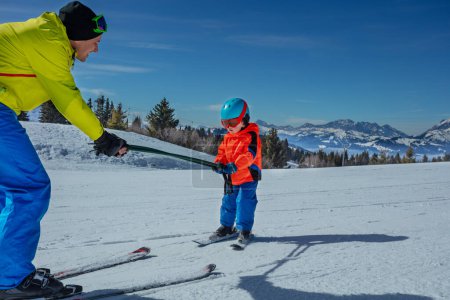 Foto de El hombre enseña a los niños a esquiar en la ladera alpina rodando por debajo y sosteniendo postes - Imagen libre de derechos