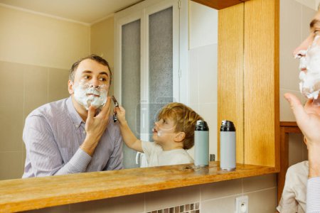 Téléchargez les photos : Père rasage regarder miroir dans la salle de bain avec enfant tenant rasoir aidant à se raser - en image libre de droit