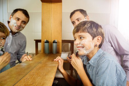 Téléchargez les photos : Sourire adolescent rasage avec père regardant dans salle de bain miroir ayant première expérience de rasage - en image libre de droit