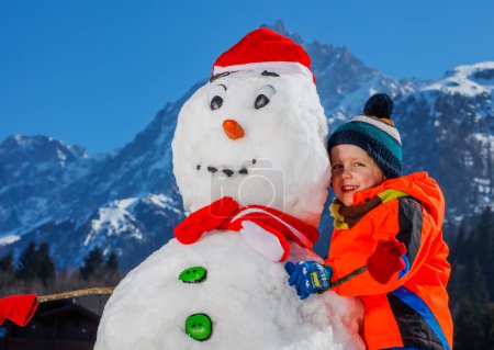 Photo for Close-up portrait of a cute little boy in winter sport outfit play with snowman in Santa hat outside - Royalty Free Image