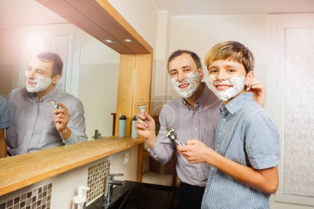 Téléchargez les photos : Papa et enfant souriant à la caméra, posant avec de la mousse sur le visage, rasoir dans les mains pendant l'adolescence premier rasage dans la salle de bain ensoleillée - en image libre de droit