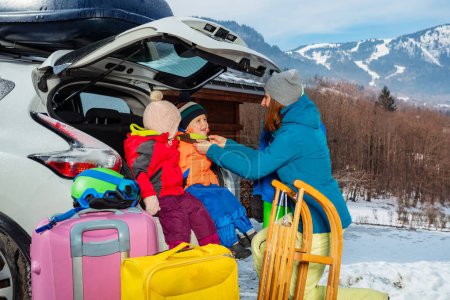 Photo for Mother help to dress kids sitting in the open trunk of the car unloading baggage sledge and bags as them arrive at winter ski snow resort - Royalty Free Image