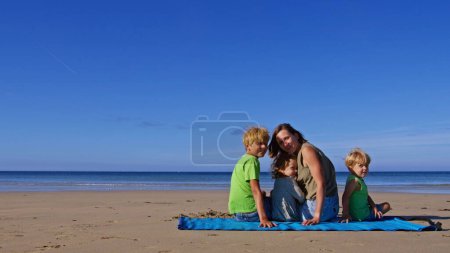 Foto de Mamá con tres niños rubios y una niña se sientan en la alfombra de la playa en el océano regresando en el día soleado - Imagen libre de derechos