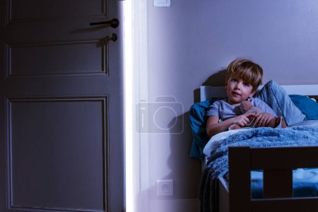 Foto de Pequeño niño tranquilo positivo sentarse en la cama sosteniendo peluche cubierto de manta cerca de la puerta ligeramente abierta con luz del día - Imagen libre de derechos