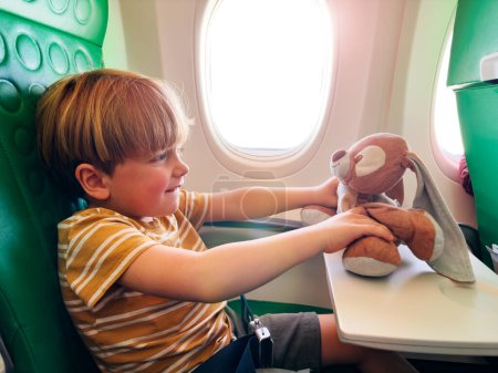 Foto de Lindo niño se sienta cerca de la ventana en el avión jugando con un juguete de peluche y sonríe en la mesa - Imagen libre de derechos
