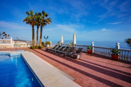 Foto de Fondo de vacaciones con chaise longs sombrillas de playa proyectando sombra cerca de la piscina y palmeras sobre el mar Mediterráneo - Imagen libre de derechos