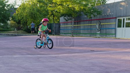Photo for Happy little boy learning to ride bicycle with extra wheels, holds steering wheel and pedals, in the skate park - Royalty Free Image