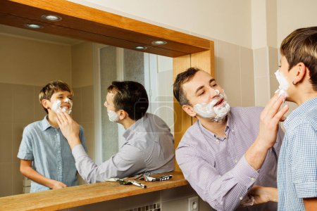 Téléchargez les photos : Papa mousse visage de l'adolescent avec sa main se préparant pour le premier rasage, souriant ensemble dans la salle de bain, vue à travers le miroir de la salle de bain - en image libre de droit