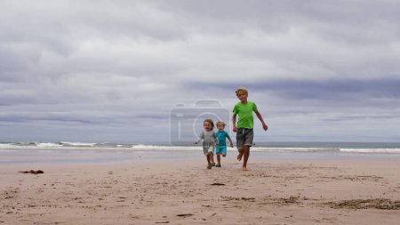Téléchargez les photos : Trois enfants, garçons et petite fille en tenue d'été courent ensemble sur la plage de sable océan souriant en s'amusant - en image libre de droit