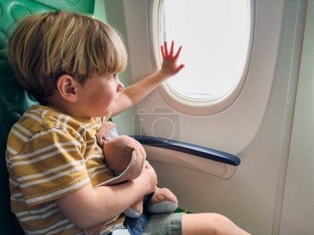 Foto de Lindo pequeño niño sentarse cerca ventana en el avión abrazo peluche juguete celebración porthole con mano - Imagen libre de derechos