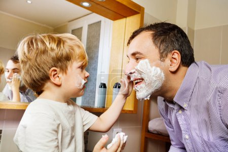 Téléchargez les photos : Petit gamin blond aidant son père à se raser le visage moussant avant le rasage du matin - en image libre de droit