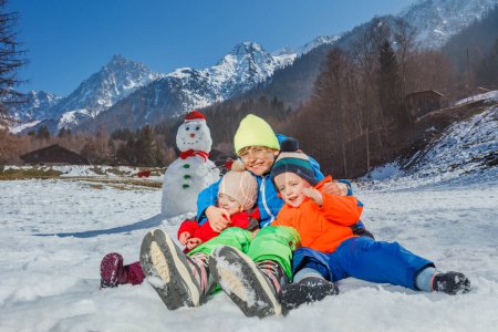 Téléchargez les photos : Là enfants heureux frères et sœurs assis ensemble près bonhomme de neige sur fond de montagnes étreignant - en image libre de droit