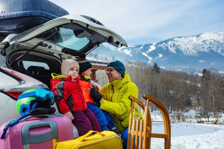 Photo for Father help to dress kids sitting in the open trunk of the car unloading baggage, sledge and bags as them arrive at winter ski snow resort - Royalty Free Image