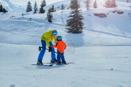 Foto de Papá en la escuela de esquí se desliza hacia atrás enseñando a los niños a esquiar yendo cuesta abajo sosteniendo los postes juntos - Imagen libre de derechos