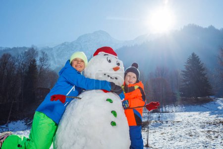 Téléchargez les photos : Faible angle clos-up photo de deux garçons heureux frères en tenue de sport d'hiver jouer avec bonhomme de neige dans le chapeau de Père Noël à l'extérieur le jour ensoleillé - en image libre de droit