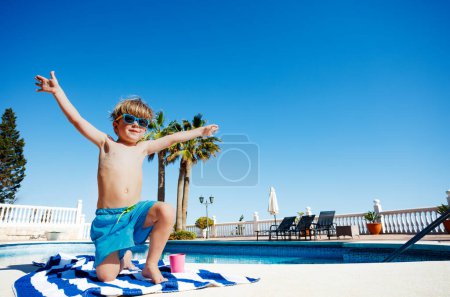 Photo for Happy kid sitting near the pool in sunglasses on striped towel raise his arms up over blue sky and palms - Royalty Free Image