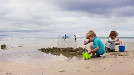 Téléchargez les photos : Deux enfants jouent avec le sable sur la plage de l'océan sur les vagues et les surfeurs passant en arrière-plan - en image libre de droit