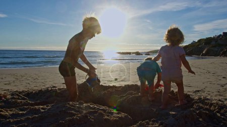 Téléchargez les photos : Groupe de trois enfants garçons et filles jouent dans le sable, creuser sur la plage à l'aide d'un seau en plastique avec des pelles - en image libre de droit