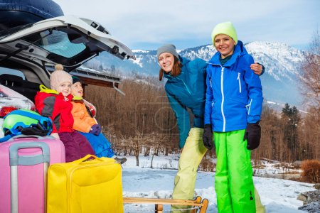 Photo for Mom stand hugging with children standing by open car trunk arrived at alpine skiing resort unloading suitcases and baggage - Royalty Free Image