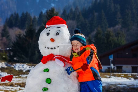 Photo for Close-up portrait of a happy little boy in winter sport outfit play with snowman in Santa hat outside - Royalty Free Image