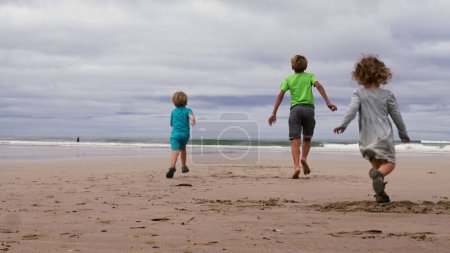 Photo for View from behind of three children, boys and little girl in summer outfit run together on the sand ocean beach having fun - Royalty Free Image