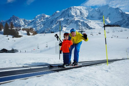 Foto de Un niño pequeño y un papá suben a la pista de esquí moviéndose cinturón en la escuela de esquí durante las vacaciones de invierno mirando a un lado, saludando la mano - Imagen libre de derechos