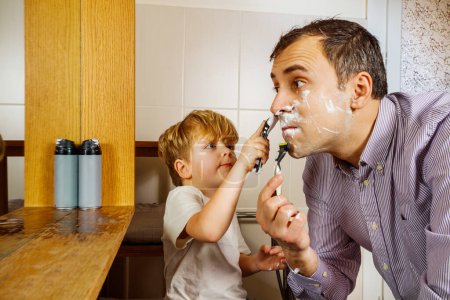 Téléchargez les photos : Petit assistant aider son père à se raser dans la salle de bain du matin avant le travail en utilisant un rasoir - en image libre de droit