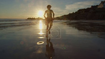 Téléchargez les photos : Un enfant en short de bain court sur la plage de l'océan éclaboussant de vagues d'eau au-dessus d'un soleil orange doux au coucher du soleil en France, Bretagne - en image libre de droit