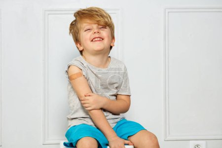 Photo for Portrait of a smiling and laughing handsome little boy with band-aid on his shoulder touching hand looking at camera - Royalty Free Image