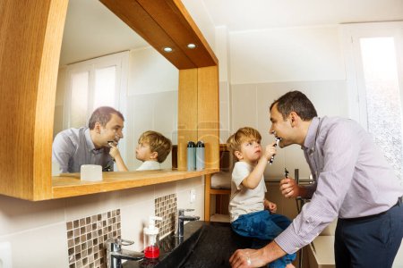 Téléchargez les photos : Père rasage et petit enfant se rasant la joue avec une lame de rasoir dans la salle de bain du matin avant le travail - en image libre de droit