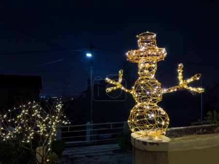 Foto de Marco iluminado muñeco de nieve de Navidad con sombrero, manos iluminación decoración por la noche sobre el cielo oscuro y luces de calle - Imagen libre de derechos