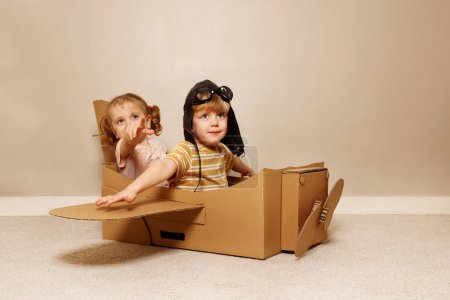 Foto de Dos niños felices niño y niña se sientan en el avión de cartón hecho a sí mismo con alas, hélice con sombrero de piloto, gafas pretenden volar - Imagen libre de derechos