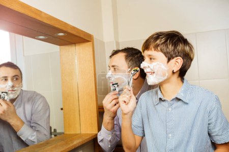 Téléchargez les photos : Père et le fils s'amusent dans la salle de bain se rasant ensemble en regardant dans le miroir souriant portant chemise - en image libre de droit