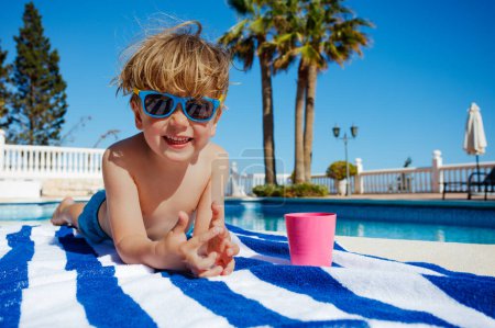 Foto de Lindo niño sonriendo y acostado en la toalla en gafas de sol cerca de la piscina en el día soleado - Imagen libre de derechos