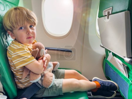 Foto de Pequeño niño sentarse junto a ojo de buey en el avión abrazando peluche juguete con la cara tranquila - Imagen libre de derechos