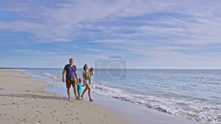 Photo for Family of four comprising a mother father little boy and girl walk, hold hands carrying their beloved sister together along the beach - Royalty Free Image