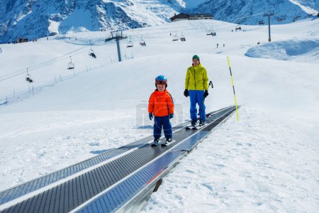 Photo for Little boy and a instructor go up on the ski moving walkway belt at skiing school during winter vacations - Royalty Free Image