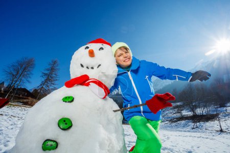 Téléchargez les photos : Mignon jeune garçon jouer câlin bonhomme de neige portant une écharpe chapeau rouge avec des mitaines sur les bâtons, debout sur les montagnes et l'éruption du soleil - en image libre de droit