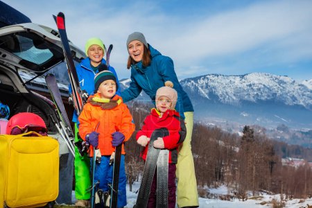 Téléchargez les photos : Mère debout tenant le ski par coffre de voiture ouvert est arrivé avec 3 enfants à la station de ski alpin déchargeant valises et bagages - en image libre de droit