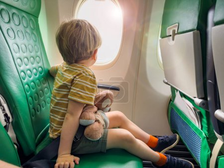 Foto de Lindo niño sentado cerca de la ventana en el avión mirando porthole durante el primer vuelo - Imagen libre de derechos