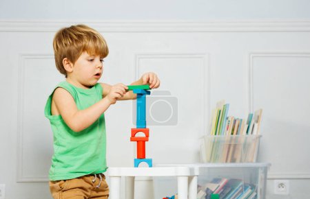 Photo for Little boy build a toy tower playing with wooden blocks sitting on the carpet in a living room over boxes of toys and books around - Royalty Free Image