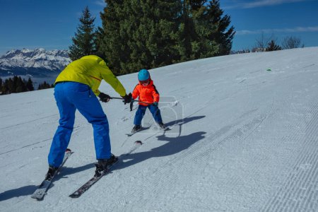 Foto de Instructor enseña a niño pequeño a esquiar en la ladera alpina rodando por debajo y sosteniendo postes - Imagen libre de derechos