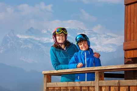 Téléchargez les photos : Mère et fils deux skieurs debout dans le balcon sur les montagnes profitant de la vue, se détendre dans l'hôtel chalet - en image libre de droit
