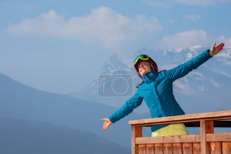 Foto de Mujer esquiadora en traje de esquí y casco de pie en el alto balcón de chalet de montaña en los Alpes, sonriendo, mirando magníficas montañas panorama - Imagen libre de derechos