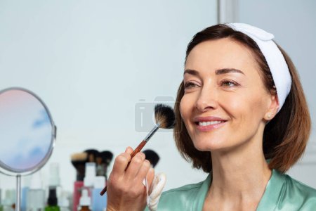 Foto de Maquillaje artista usando cepillo de ventilador en la cara de la mujer de mediana edad con diadema sentado ante el espejo - Imagen libre de derechos