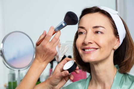 Photo for Close-up portrait of a wide smiling middle aged woman model with beautician applying powder using large brush - Royalty Free Image