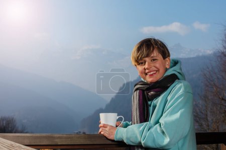 Téléchargez les photos : Joyeux adolescent garçon snowboarder debout avec tasse de café portant écharpe, sweat-shirt, sur le balcon en bois du chalet et admire les montagnes alpines ensoleillées, les forêts - en image libre de droit