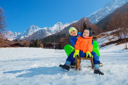 Téléchargez les photos : Deux garçons souriants heureux, frères assis sur le traîneau descendant la pente de la montagne - en image libre de droit