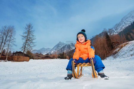 Téléchargez les photos : Garçon en tenue d'hiver assis sur le traîneau sur le point de glisser descente souriant et avoir du plaisir en vacances - en image libre de droit